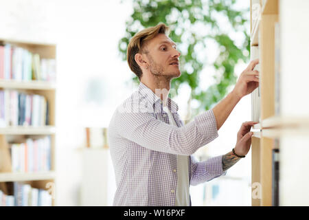 Ernsthafte konzentriert hübscher junger Mann mit Stoppeln am Bücherregal und finden Buch im Regal in der Bibliothek Stockfoto