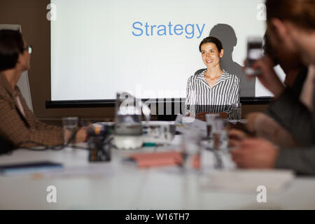 Fröhlich zuversichtlich attraktive junge weibliche Manager in abgestreift Bluse sitzen gegen Projektionsfläche und Strategie zu Kollegen an der Konferenz Stockfoto
