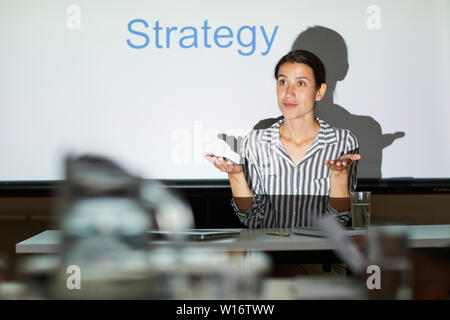 Inhalt selbstbewussten jungen Dame in abgestreift Bluse sitzt am Tisch gegen Projektionsfläche in dunklen Zimmer und diskutieren Strategie für die Verwirklichung der Ziel wit Stockfoto