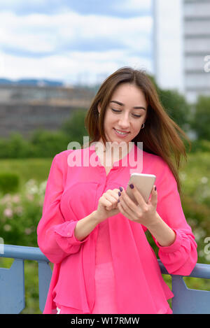 Junge Frau Wählen einer Nummer auf Ihrem Handy mit einem glücklichen Lächeln der Vorfreude, als Sie auf einer Brücke, lehnte sich an die Balustrade steht Stockfoto