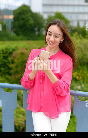 Junge Frau liest eine SMS auf Ihr Handy mit einem glücklichen Lächeln, als sie draußen an einem metallgeländer mit Blick auf einen Garten steht Stockfoto