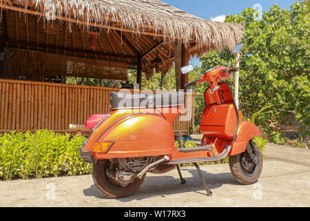 Eine alte orange und schwarz Vespa Super motorscooter ist auf einem Bürgersteig geparkt. Rückansicht eines hellen orange motor scooter außerhalb eines Bambus Haus geparkt Stockfoto