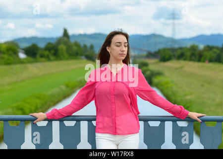 Stilvolle junge Frau steht auf einer Brücke denken ihre Arme ruht auf dem Geländer und geschlossenen Augen mit einem nachdenklich ernsten Ausdruck gegen eine ländliche l Stockfoto
