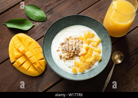 Natürliche Joghurt mit Mango slices und crunchy Müsli in der Schüssel und ein Glas Orangensaft auf einem Holztisch Hintergrund. Ansicht von oben. Sauber Essen, Diäten, Stockfoto