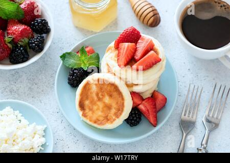 Ferienhaus syrniki mit Erdbeeren, Honig auf blauem Teller und Tasse schwarzen Kaffee für den Hintergrund. Gesund leckeres Frühstück Stockfoto