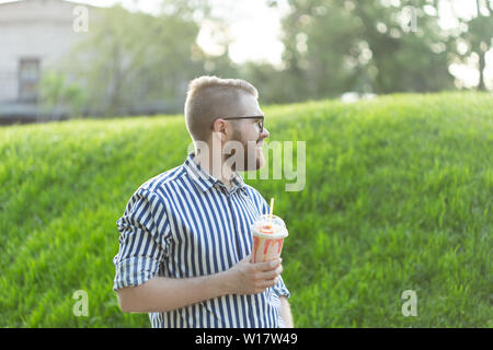 Seitenansicht eines stilvollen junger Mann mit einem Bart Holding einen Milchshake und bewundern Sie die Aussicht auf die Stadt zu Fuß in den Park an einem warmen Sommertag. Das Konzept der Stockfoto