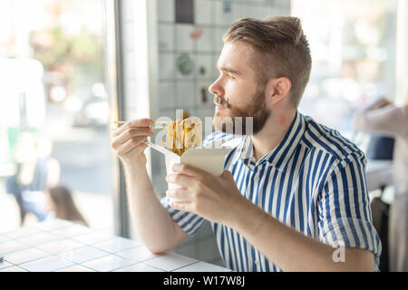 Seitenansicht eines hübschen Jungen hipster Kerl essen Udon Nudeln mit hölzernen Eßstäbchen in einem Cafe saßen und in das Fenster. Asiatische Küche Stockfoto