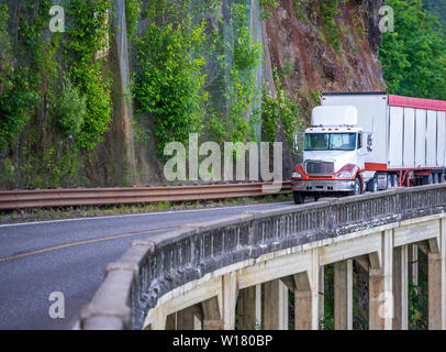 Big Rig tag Fahrerhaus mit dachspoiler Semi Truck Transport von kommerziellen Ladung im abgedeckten bulk Auflieger Fahren auf schmalen Straßen mit Felswand Stockfoto