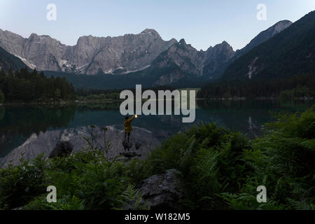 Junge in gelb Regenjacke stehend auf dem Rock und halten eine leuchtende Hand Laterne an der szenischen Sommer See Landschaft in der Dämmerung, Laghi di Fusine, Italien Stockfoto