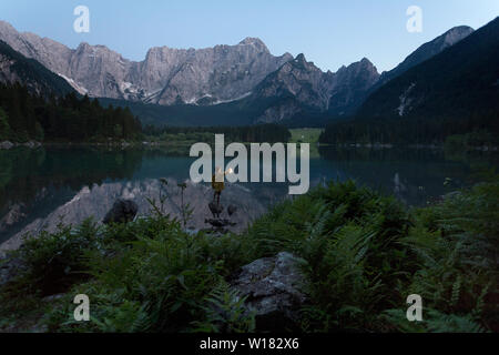 Junge in gelb Regenjacke stehend auf dem Rock und halten eine leuchtende Hand Laterne an der szenischen Sommer See Landschaft in der Dämmerung, Laghi di Fusine, Italien Stockfoto