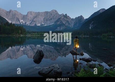 Junge in gelb Regenjacke stehend auf dem Rock und halten eine leuchtende Hand Laterne an der szenischen Sommer See Landschaft in der Dämmerung, Laghi di Fusine, Italien Stockfoto