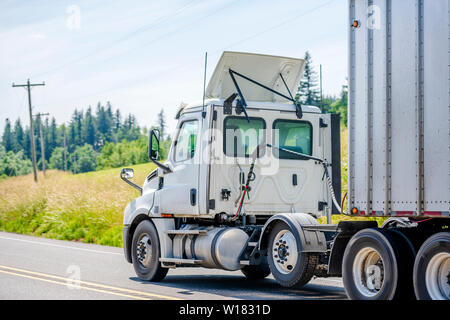 Big Rig White Day Cab Semi Truck mit Dachspoiler transportieren riesige überdachte bulk Auflieger abwärts bewegen auf dem drehen, kurvenreiche Straße mit gelben Stockfoto