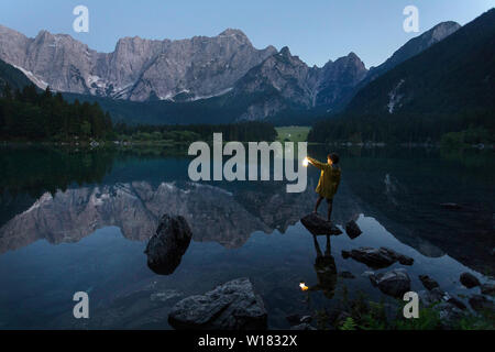 Junge in gelb Regenjacke stehend auf dem Rock und halten eine leuchtende Hand Laterne an der szenischen Sommer See Landschaft in der Dämmerung, Laghi di Fusine, Italien Stockfoto