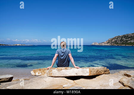 Mann sitzt auf einem Felsen und genießen Sie den wunderschönen Ausblick auf das Meer an einem Strand mit einem klaren blauen Himmel in Sardinien (Italien) Stockfoto