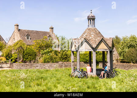 Maytime in den Cotswolds - Zwei weibliche Radfahrer in der 1874 Pumpenhaus von alten Steinhäusern mit Blick auf das Dorf grün im Dorf von Farmington UK Stockfoto