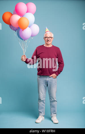 Gerne reifer Mann in Geburtstag Gap und Casualwear holding Ballons während auf blauem Hintergrund posiert Stockfoto