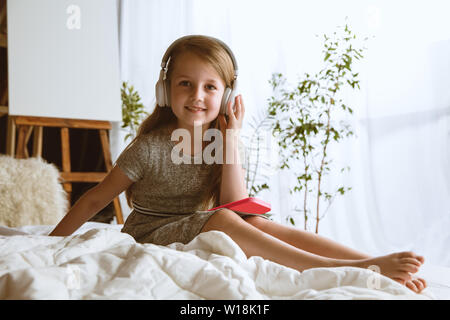 Mädchen über verschiedene Geräte zu Hause. Kleines Modell in ihrem Bett sitzt mit großen Kopfhörer Anhören von Musik und zum Genießen. Konzept der Interaktion von Kindern und moderne Technologien. Stockfoto