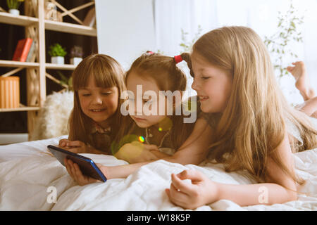 Mädchen über verschiedene Geräte zu Hause. Childs mit Smartphone liegen und lächelnd, Videos oder Spiele spielen. Die selfie, chatten, spielen. Interaktion von Kindern und moderne Technologien. Stockfoto