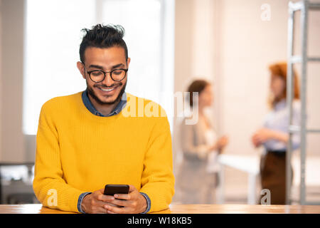 Positive hübscher junger arabischer Manager in Gläsern an der Theke stehend in Büro- und Simsen sms auf Telefon Stockfoto