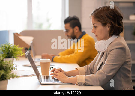 Ernsthafte konzentrierte junge Frau mit Kopfhörer auf dem Hals sitzen am Tisch und Tippen auf Laptop im Büro Stockfoto
