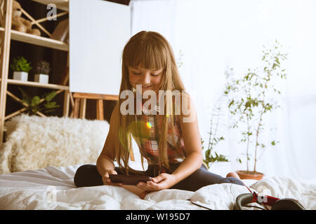 Mädchen über verschiedene Geräte zu Hause. Kleines Modell in ihrem Bett sitzend mit Smartphone und selfie oder über Videochat mit ihren Freunden. Konzept der Interaktion von Kindern und moderne Technologien. Stockfoto