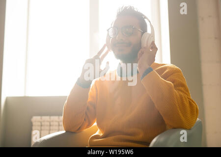Inhalt sorglos hipster Kerl in drahtlosen Kopfhörern und Brillen in einem Sessel sitzend vor Sonnenlicht und genießen Sie entspannende Musik Stockfoto
