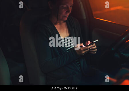 Frau SMS auf Handy im Auto in der Nacht auf einem Parkplatz, Erwachsene weibliche Person mit Smartphone für Kommunikation, selektiven Fokus Stockfoto