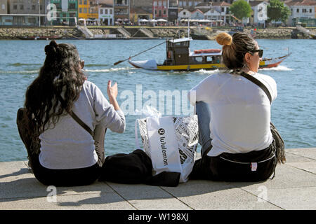 Zwei Frauen Rückansicht zurück hinter das Sitzen auf dem Kai mit lululemon Beutel auf dem Fluss Douro in Ribeira Porto Oporto Portugal Europa EU-KATHY DEWITT Stockfoto