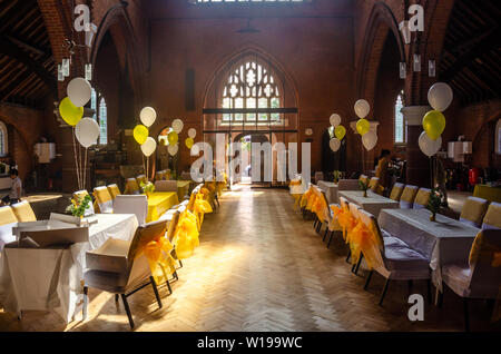 Eine Kirche Halle mit Tischen und Stühlen im weißen Stoff mit gelbem Band Bogen bereit für eine Party oder Rezeption gekleidet. Stockfoto