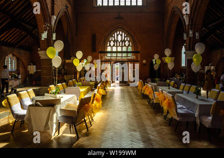 Eine Kirche Halle mit Tischen und Stühlen im weißen Stoff mit gelbem Band Bogen bereit für eine Party oder Rezeption gekleidet. Stockfoto