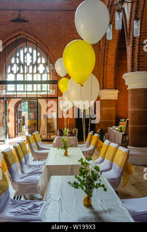 Tische und Stühle in einer Kirche Halle, mit Helium gefüllte Ballons und gelben Bändern bereit für eine Party, Empfang oder Feier eingerichtet Stockfoto