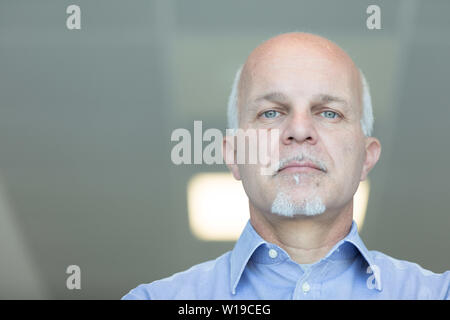 Senior balding Mann mit blauen Augen und einem ausdruckslosen Ausdrucks nach unten in die Kamera starrt in einem niedrigen Winkel Kopf Porträt im Innenbereich Stockfoto
