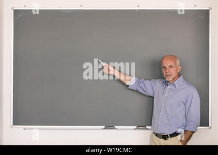 Senior Lehren stehen mit ernstem Gesichtsausdruck und mit der Hand in der Tasche halten, Kreide, die auf eine leere Tafel in einem Klassenzimmer Stockfoto