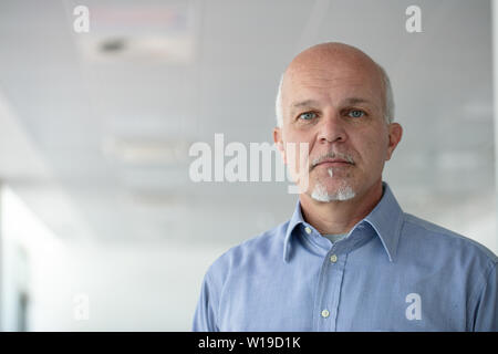 Ernsthafte älterer Mann aufmerksam in die Kamera starrte mit einem durchdringenden Blick in einem niedrigen Winkel anzeigen gegen eine weiße Decke mit Kopie Raum Stockfoto