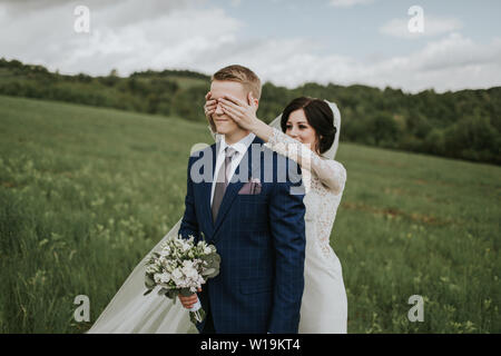 Braut, die Augen des Bräutigams. Fröhliche Bräutigam stehen auf Wiese und Braut, die seine Augen von hinten. Stockfoto