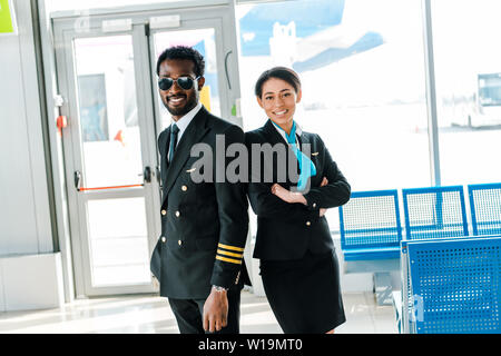 Lächelnd afrikanische amerikanische Piloten Sonnenbrille und stewardess mit verschränkten Armen gemeinsam im Flughafen Stockfoto