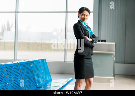 Lächelnd afrikanische amerikanische Stewardess Stehen mit verschränkten Armen im Flughafen und auf der Suche an Kamera Stockfoto