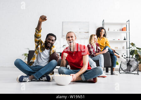 Fröhlicher und positiver multikulturelle junge Leute sitzen auf dem Boden und beobachten Sport Spiel mit Schüssel Popcorn Stockfoto