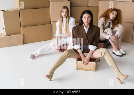 Schöne multikulturelle Mädchen sitzen auf Koffern in der Nähe der Boxen auf Weiß Stockfoto