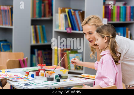 Attraktiven Lehrer in der Nähe von cute kid Malerei auf Papier in Bibliothek Stockfoto