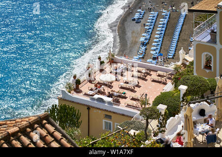 Super sonnig Stadtbild von Positano Stadt an der Amalfiküste in Italien. Es ist ein Strand mit vielen blauen Schirme und Sonnenliegen, Häuser mit Terrassen mit ch Stockfoto