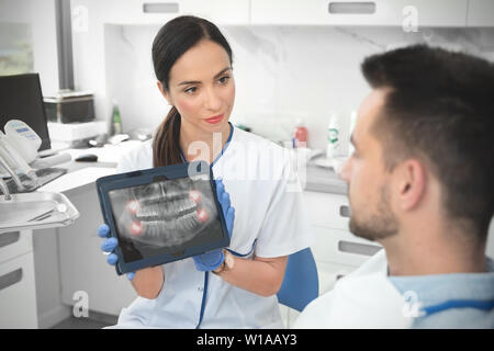 Weiblichen Zahnarzt zeigt Zähne x-ray auf digitalen tablet Bildschirm. Patient sitzt auf Stuhl in professionelle zahnmedizinische Klinik. Stockfoto