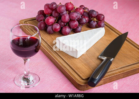 Glas Rotwein und ein dreieckiges Stück Brie Käse, roten süßen Trauben, Kochmesser auf braunem Holz Schneidebrett. Frischkäse mit genießbaren Schimmel Stockfoto