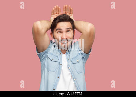 Portrait von Lustig verrückt gut aussehender bärtiger junger Mann in blau casual style Hemd ständigen bunny Geste die Hände auf den Kopf und suchen. indoor Studio shot, ist Stockfoto