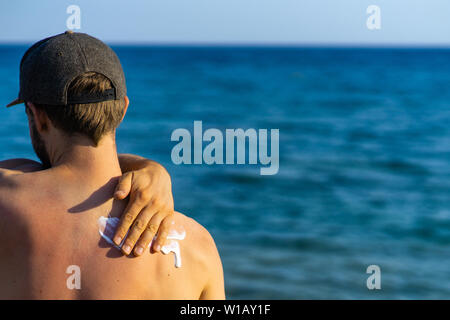 Junger Mann mit Wave-gebildet Sonnencreme auf seiner Schulter stehen am Strand, über dem Meer Stockfoto