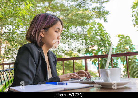 Frau im schwarzen Anzug Eingabe laptop neben Tasse Kaffee Stockfoto