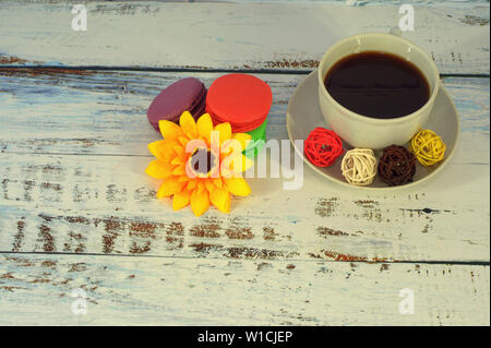 Vier Makronen in verschiedenen Farben mit einem blütenknospe eingerichtet liegen auf einem Tisch neben einer Tasse Kaffee auf eine Untertasse mit einem Löffel. Stockfoto