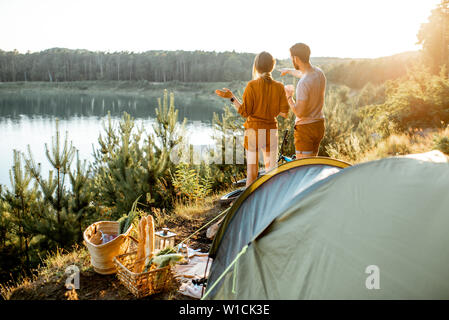 Junges Paar stehend auf dem Campingplatz, genießen Sie einen wunderschönen Blick auf den See, unterwegs in den Bergen auf den Sonnenuntergang Stockfoto