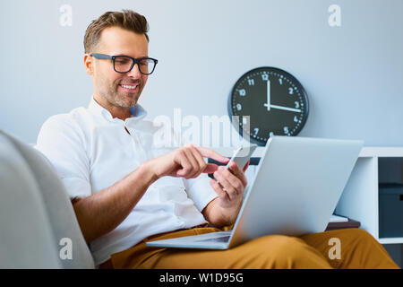 Junger Mann mit seinem Smartphone für das Online Banking - Sitzen auf einem Sofa mit Laptop auf dem Sprung Stockfoto
