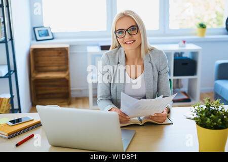 Junge Geschäftsfrau auf Kamera, während im Home Office arbeiten mit Laptop und Dokumente Stockfoto